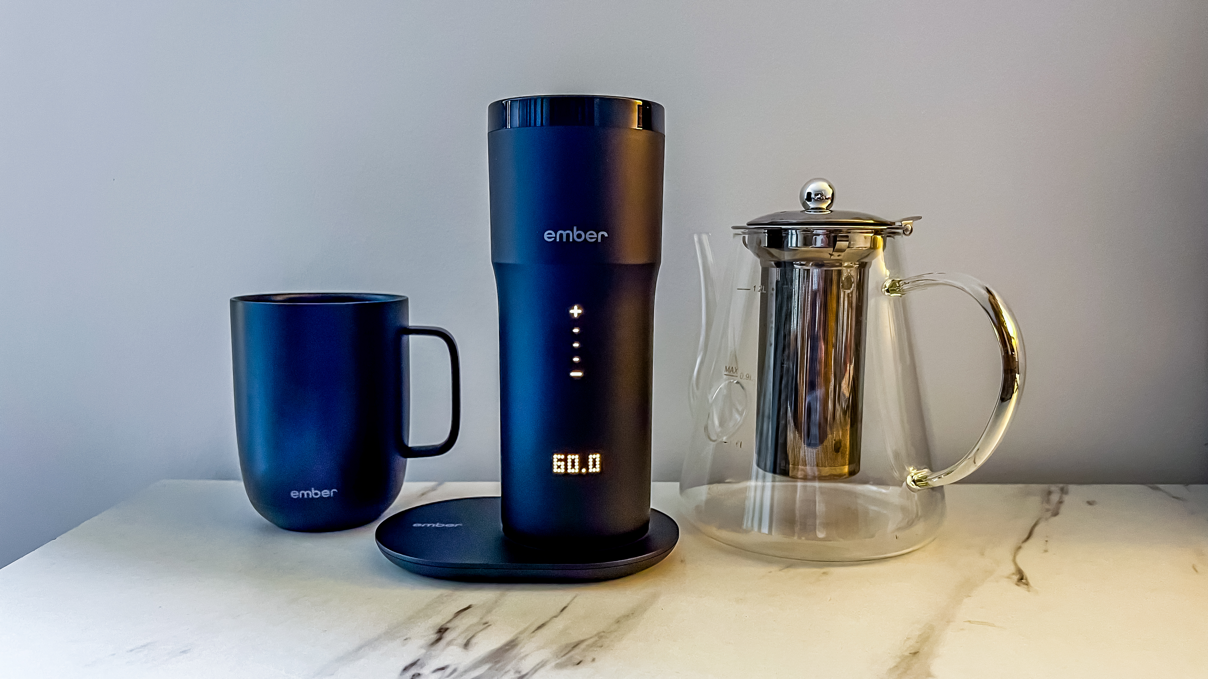 Ember Travel Mug 2+ with lit display on its charging plate sitting between the Ember Mug 2 and a glass kettle